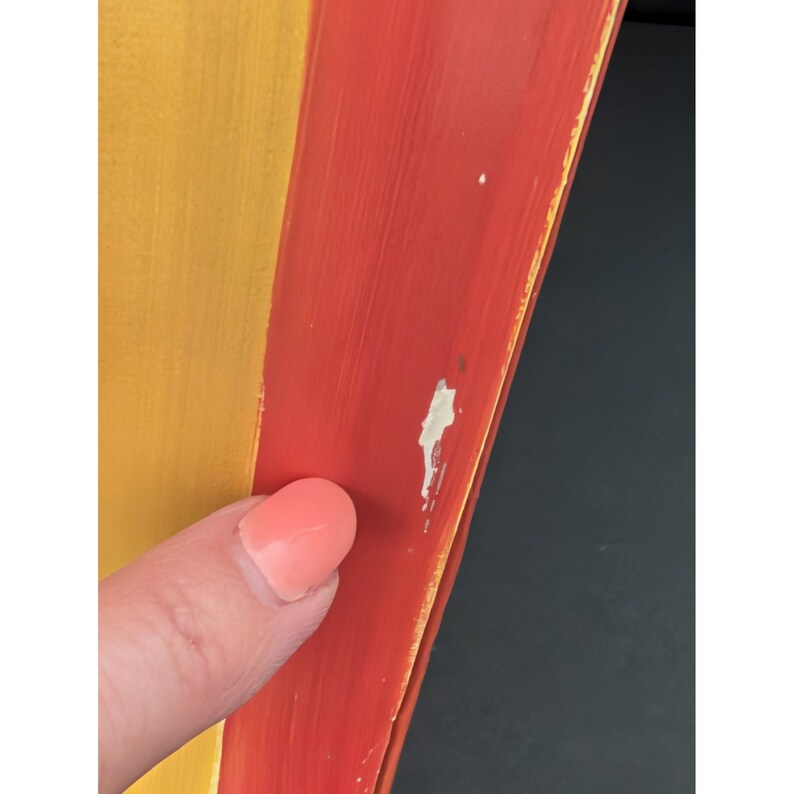 May include: Close-up of a section of a painted wooden object. The object is divided into two sections, one yellow and one red. The red section has some paint chipping. A finger with pink nail polish is touching the object.