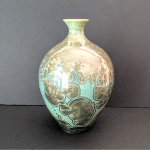 May include: A decorative ceramic vase with a unique, textured pattern. The vase features a light green rim and base, with a turquoise and brown mottled design covering the body. The vase has a rounded shape and a narrow neck.