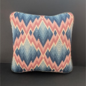 May include: Square decorative pillow with a geometric pattern in shades of blue, pink, and cream. The design features a repeating diamond motif. The pillow has a dark blue border and is likely made of a woven fabric.