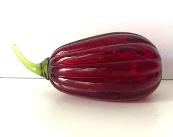 May include: A decorative glass object shaped like a red pepper. The pepper has a ribbed texture and a green stem. The glass is a deep, translucent red color, and the stem is a bright green.