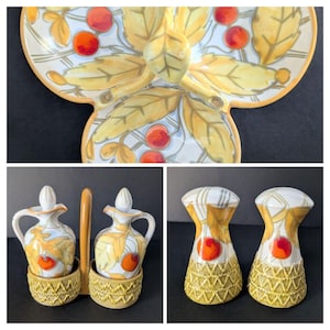 May include: A ceramic serving set with a floral design featuring yellow leaves and red fruit. The set includes a decorative plate, two oil and vinegar cruets with a holder, and two salt and pepper shakers. The pieces have a cream and yellow color scheme.