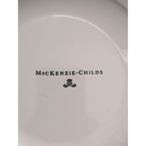 May include: White ceramic plate with the text "MacKenzie-Childs" in dark gray, along with a thistle emblem. The plate has a slightly curved rim and a smooth surface.