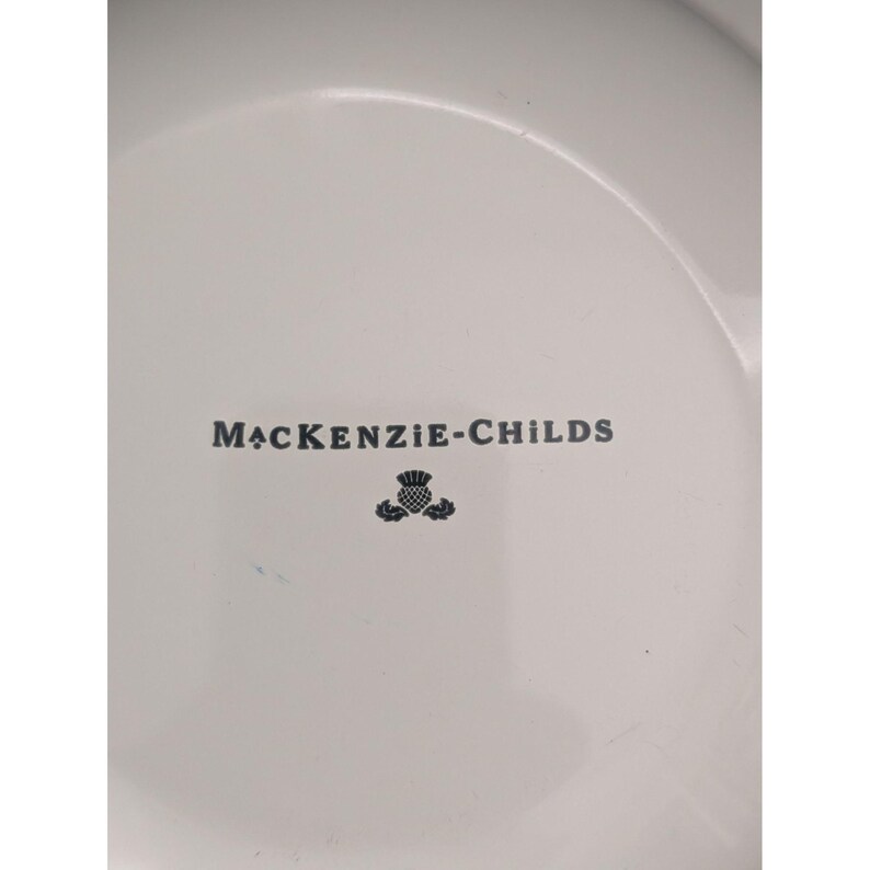 May include: White ceramic plate with the text "MacKenzie-Childs" in dark gray, along with a thistle emblem. The plate has a slightly curved rim and a smooth surface.