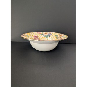 May include: A white ceramic bowl with a wide, floral-patterned rim. The rim features a gold edge and a design of yellow polka dots and colorful flowers. The bowl's interior is white, and the overall design is elegant.