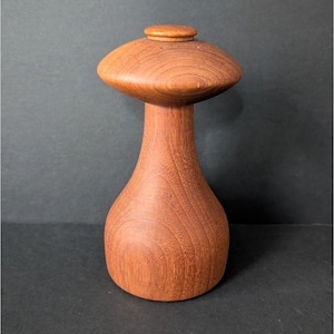 Może przedstawiać: Drewniany młynek do pieprzu z grzybkowatą górą i pękatą podstawą. Widoczne usłojenie drewna, kolor ciepły czerwonobrązowy. Młynek to przyrząd kuchenny.