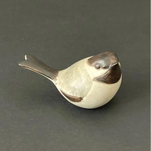May include: A ceramic bird figurine with a light gray body and a dark brown head, tail, and wing accents. The bird is in a resting pose, with a rounded body and a detailed beak. The figurine is a decorative item.