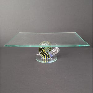 May include: A clear glass serving tray with a rectangular top. The tray is supported by a snail-shaped base with yellow and black stripes. The base sits on a round, clear glass foot.