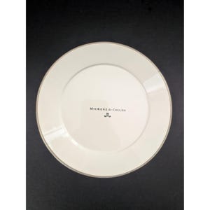 Op de afbeelding: Witte keramische plaat met een gouden rand. De plaat heeft de tekst "MacKenzie-Childs" in zwart gedrukt. De plaat is rond en heeft een eenvoudig, elegant ontwerp.