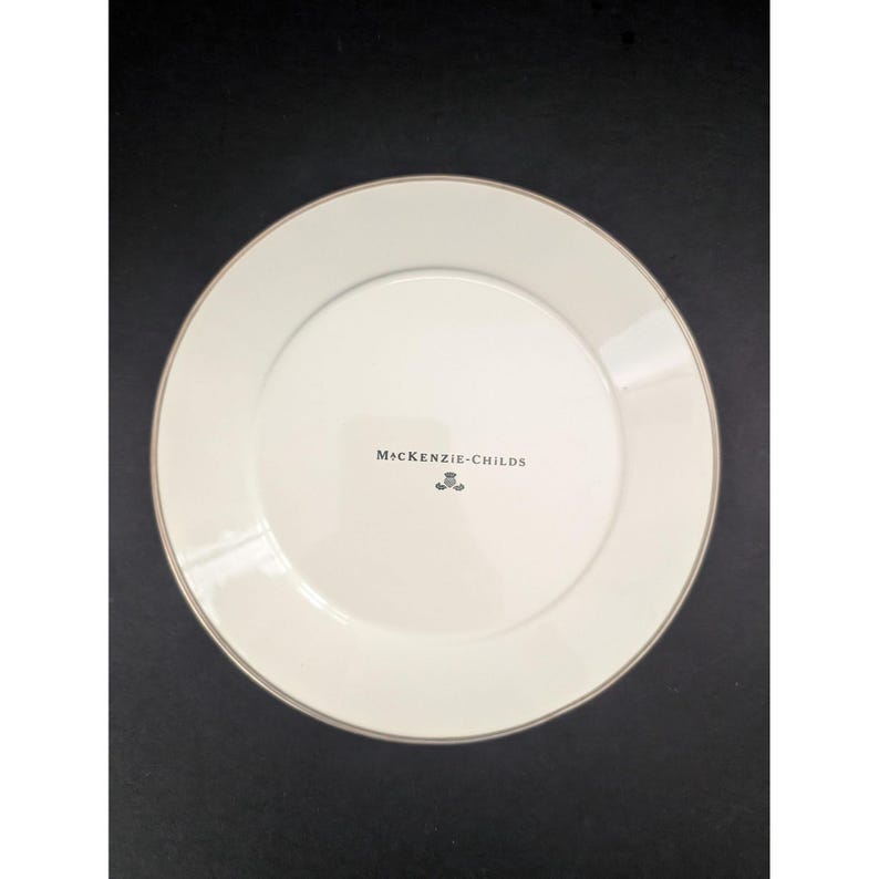Op de afbeelding: Witte keramische plaat met een gouden rand. De plaat heeft de tekst "MacKenzie-Childs" in zwart gedrukt. De plaat is rond en heeft een eenvoudig, elegant ontwerp.