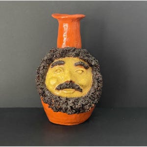 Könnte beinhalten: Eine handgefertigte Keramikvase mit einem einzigartigen Gesichtsdesign. Die Vase hat ein gelbes Gesicht mit einem schwarzen Schnurrbart und Bart, vor einem orangefarbenen Sockel und Hals. Der strukturierte Bart verleiht diesem Dekorationsstück eine besondere Note.