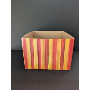 May include: A rectangular cardboard box with vertical stripes in shades of red, orange, and yellow. The box has a plain, unpainted interior and sits on a dark surface. The stripes are hand-painted, giving the box a unique, artistic look.