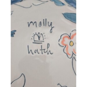 May include: White ceramic plate with hand-painted floral and leaf designs in blue, peach, and yellow. The plate features the text "molly hatch" in dark blue script, along with a crown symbol.
