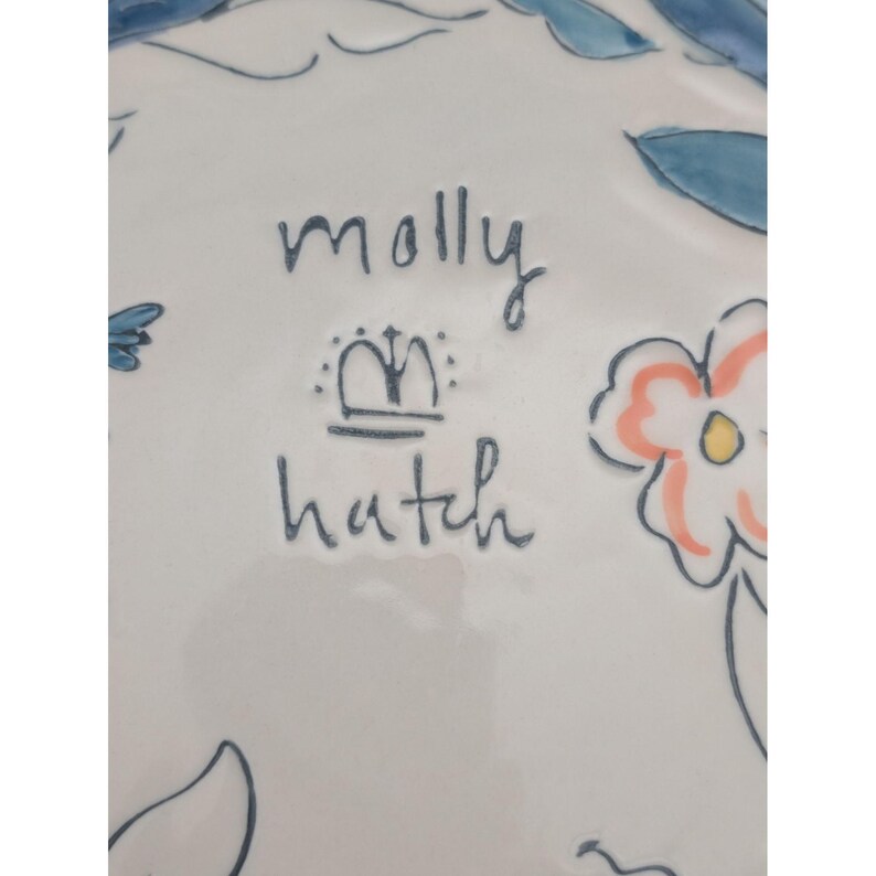 May include: White ceramic plate with hand-painted floral and leaf designs in blue, peach, and yellow. The plate features the text "molly hatch" in dark blue script, along with a crown symbol.