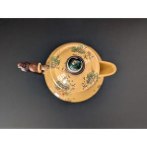 May include: Overhead view of a yellow teapot with a matching lid and saucer. The teapot features a floral pattern in green and purple. The handle is made of wood. The spout is on the right side.
