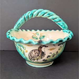 Könnte beinhalten: Eine weiße Keramikschale mit einem türkisfarbenen, gedrehten Griff. Die Schale hat einen gewellten Rand und ist mit einem gemalten Design eines Vogels und grüner Blätter verziert.