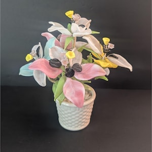 Könnte beinhalten: Ein farbenfrohes Glasblumenarrangement in einem weißen, gewebten Topf. Die Blumen haben rosa, weiße, gelbe und blaue Blütenblätter mit grünen Blättern. Das Arrangement ist vor einem dunklen Hintergrund platziert, der die leuchtenden Farben der Glasblumen hervorhebt.