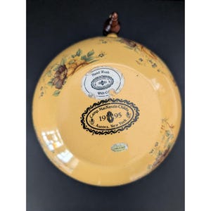 May include: A yellow decorative plate with floral and leaf designs. The plate has a brown handle and several stickers with text, including "Camp MacKenzie-Childs 1995 Aurora, New York."