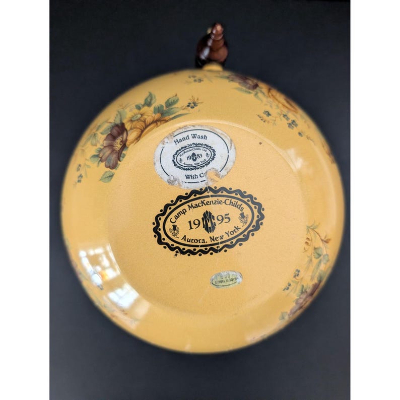 May include: A yellow decorative plate with floral and leaf designs. The plate has a brown handle and several stickers with text, including "Camp MacKenzie-Childs 1995 Aurora, New York."