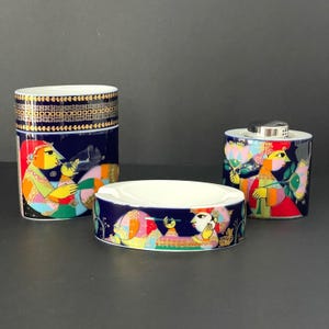 Pode incluir: Um conjunto de três peças de cerâmica com fundo azul escuro e ilustrações coloridas. O conjunto inclui um recipiente cilíndrico alto, um cinzeiro pequeno e um recipiente pequeno com tampa prateada. As ilustrações apresentam figuras em cores vibrantes.