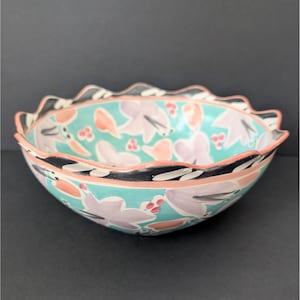 May include: A large, scalloped ceramic bowl with a turquoise glaze. The bowl is decorated with a floral pattern in shades of pink, purple, and black.