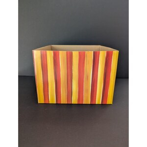 May include: A rectangular storage box with a hand-painted design of vertical stripes in shades of yellow, orange, and red. The interior and top edge of the box are a natural cardboard color. The box is empty.