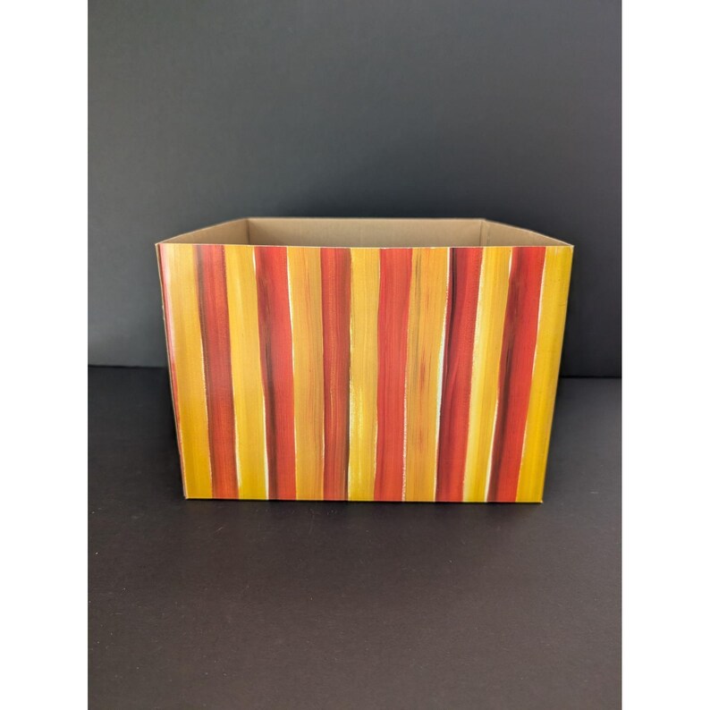 May include: A rectangular storage box with a hand-painted design of vertical stripes in shades of yellow, orange, and red. The interior and top edge of the box are a natural cardboard color. The box is empty.