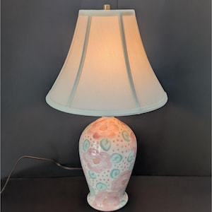 May include: A ceramic table lamp with a white lampshade and a floral design. The base is painted with pink and blue flowers and green leaves. The lamp has a gold-colored finial and a brown cord.