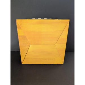 May include: A square, yellow box with a textured, painted finish. The box has a geometric design with angled sides and a black and white striped handle. The background is black.