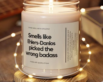 May include: A lit candle in a glass jar with a label that reads "Smells like Ehlers-Danlos picked the wrong badass." The candle sits on a round wooden coaster. The background includes a guitar and a book.