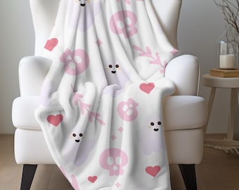 May include: A white blanket draped over a white armchair. The blanket features a repeating pattern of pink skulls, hearts, ghosts, and floral elements. The blanket is soft and plush, perfect for snuggling up on a chilly day.