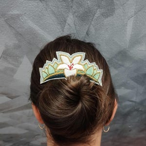 May include: A white and green floral hair accessory, embroidered with gold and red accents, is worn in a bun. The accessory has a crown-like shape, with a central flower design and a scalloped edge. The background is a gray wall.