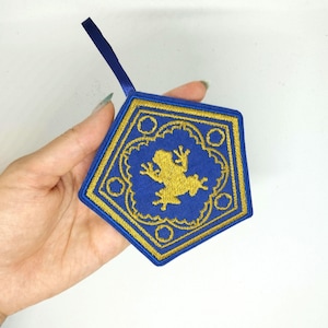 Può includere: Un ornamento ricamato blu e oro a forma di esagono con una rana all'interno. L'ornamento ha un anello per appenderlo.