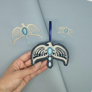 May include: Embroidered eagle ornaments in cream and navy blue, featuring a large oval blue gem at the center and smaller gems below. The ornaments have a detailed wing design and a blue ribbon for hanging. The background is a light blue.