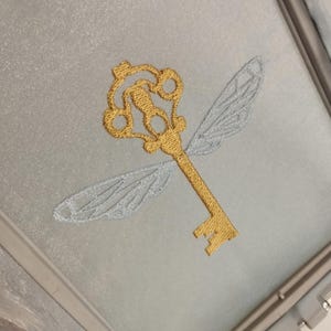 Puede incluir: Una llave dorada con alas, un símbolo popular de la serie Harry Potter, está bordada en una tela azul claro.