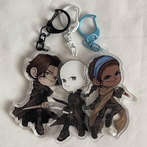 May include: Three cartoon character keychains with black, white, and brown details. The characters are a young man with brown hair, a bald man, and a young woman with brown hair and a blue headband. The keychains are attached to silver, black, and blue key rings.