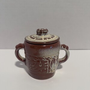 May include: A brown ceramic sugar bowl with two side handles and a matching lid. The exterior features a textured, embossed leaf pattern around the center, accented with a cream-colored glaze that fades into the dark brown base and lid top.