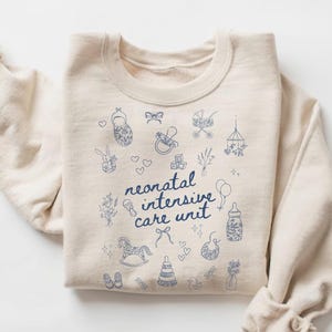 K&ouml;nnte beinhalten: Ein beiger Pullover mit einem blauen Design aus babybezogenen Symbolen und dem Text "neonatal intensive care unit".