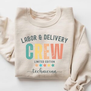 May include: A beige sweatshirt with the text "Labor & Delivery Crew Limited Edition Technician" in colorful retro-style lettering. The text is surrounded by five stars in different colors.