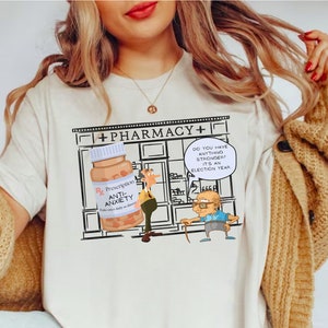 May include: White t-shirt with a cartoon illustration of a pharmacy with a pharmacist and a customer. The customer is asking for "anything stronger? It's an election year." The pharmacy is selling "Anti-Anxiety" pills.