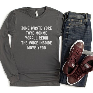 May include: A charcoal gray long-sleeve t-shirt with white text that reads "Jone Waste Yore Toye Monme Yorall Rediii The Voice Insoide Moye Yedd". The shirt is paired with dark blue jeans and maroon high-top sneakers.