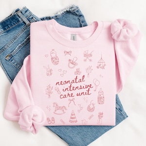 K&ouml;nnte beinhalten: Rosafarbener Pullover mit dem Text "neonatal intensive care unit" in roten Buchstaben. Der Pullover ist mit kleinen, rosafarbenen Illustrationen von Babyartikeln wie einem Schaukelpferd, einer Rassel und einem Schnuller verziert.