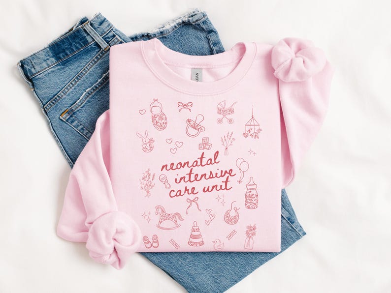 K&ouml;nnte beinhalten: Rosafarbener Pullover mit dem Text "neonatal intensive care unit" in roten Buchstaben. Der Pullover ist mit kleinen, rosafarbenen Illustrationen von Babyartikeln wie einem Schaukelpferd, einer Rassel und einem Schnuller verziert.