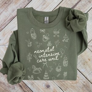 K&ouml;nnte beinhalten: Ein gr&uuml;nes Sweatshirt mit einem wei&szlig;en Design, das "neonatal intensive care unit" sagt, umgeben von kleinen Illustrationen von Babyartikeln wie einem Schaukelpferd, einer Rassel und einer Babyflasche.