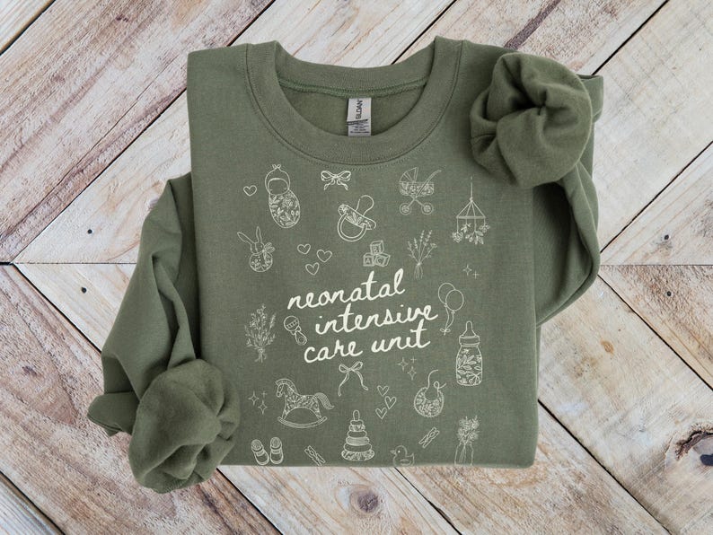 K&ouml;nnte beinhalten: Ein gr&uuml;nes Sweatshirt mit einem wei&szlig;en Design, das "neonatal intensive care unit" sagt, umgeben von kleinen Illustrationen von Babyartikeln wie einem Schaukelpferd, einer Rassel und einer Babyflasche.