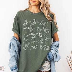 May include: Moss green t-shirt with a white graphic of cartoon geese and the text "silly goose nicu club". The shirt has short sleeves and a relaxed fit. A denim jacket is draped over the shoulders.