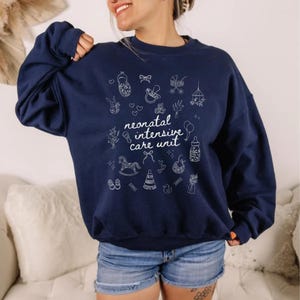 K&ouml;nnte beinhalten: Ein marineblauer Pullover mit einem wei&szlig;en Grafikdesign aus babybezogenen Symbolen und dem Text "neonatal intensive care unit".