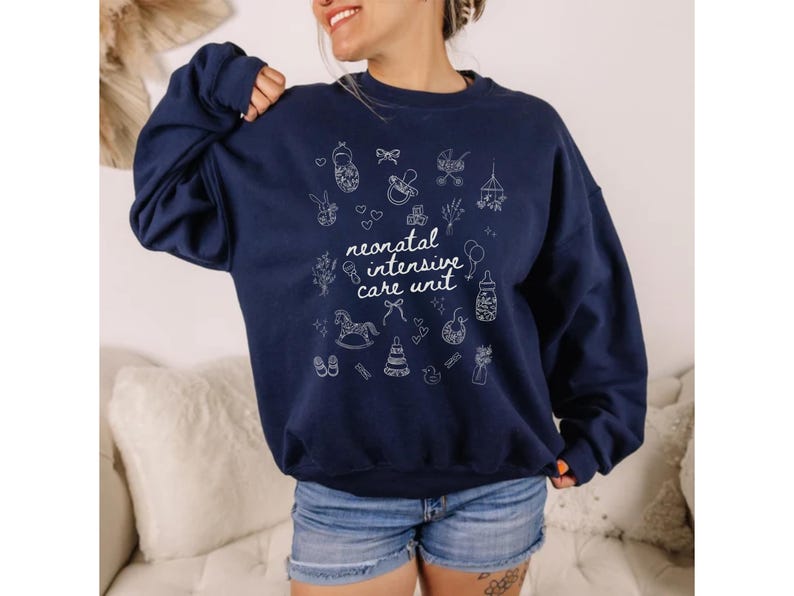 K&ouml;nnte beinhalten: Ein marineblauer Pullover mit einem wei&szlig;en Grafikdesign aus babybezogenen Symbolen und dem Text "neonatal intensive care unit".