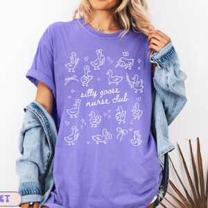 May include: Violet-colored t-shirt with a white graphic of cartoon geese and the text "silly goose nurse club." The shirt has short sleeves and a relaxed fit. A denim jacket is worn over the shirt.