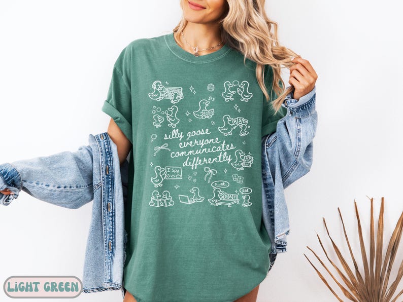 May include: Light green t-shirt with white graphic illustrations of ducks and the text "silly goose everyone communicates differently." The shirt is a relaxed fit and has rolled-up sleeves. A light blue denim jacket is worn over the shirt.