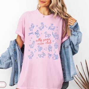 May include: A light pink t-shirt with a blue and red graphic of cartoon geese and the text "silly goose nicu club". The geese are in various poses, some wearing hats and bows. The shirt is made of soft material.
